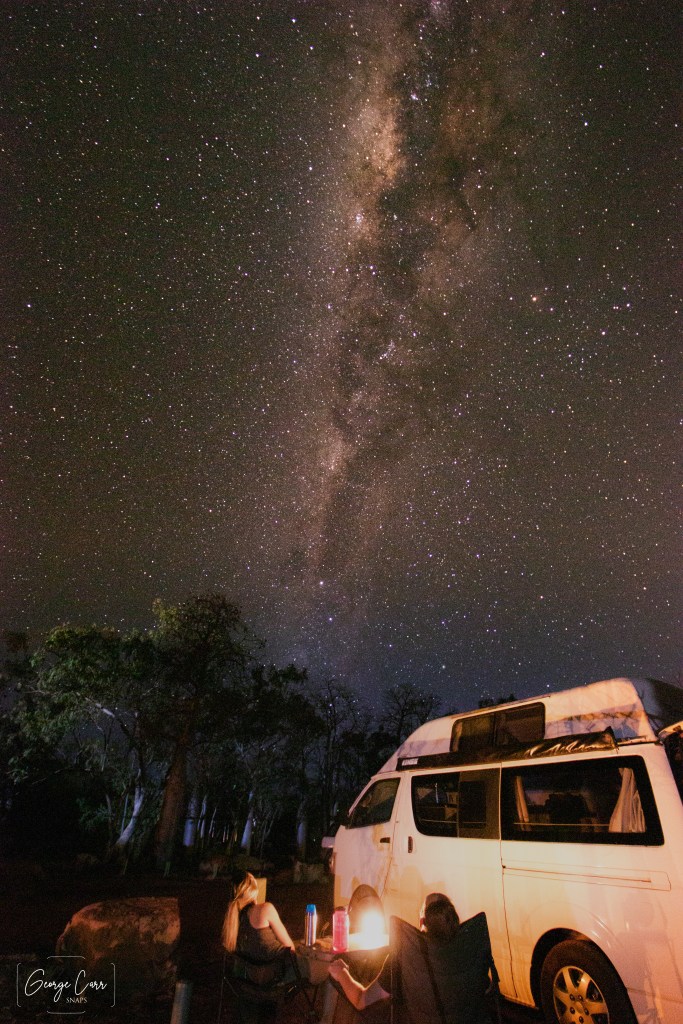 Evening image of the milky way above with two campers staring at the sky from outside their van